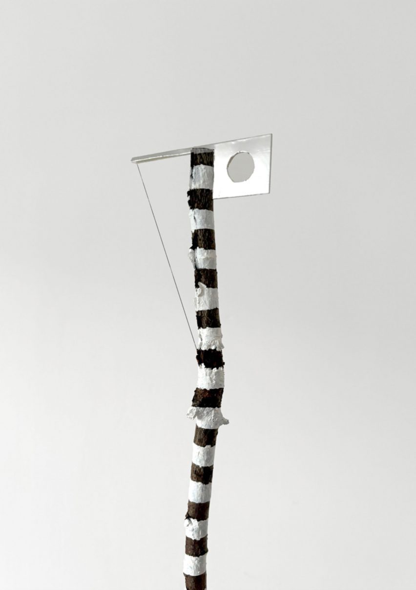 A photograph of an installation that takes form as a stick in brown and white tones, with a square silver attachment at the top.