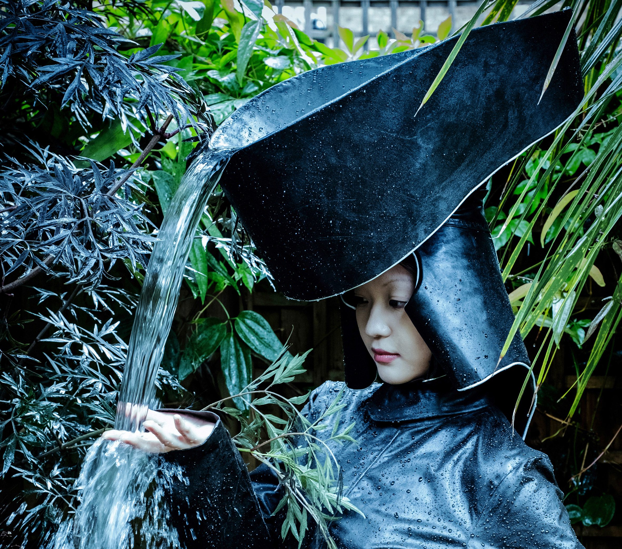 A photograph of a person wearing a large headpiece which is designed to collect rainwater, with water pouring out of it at an angle.