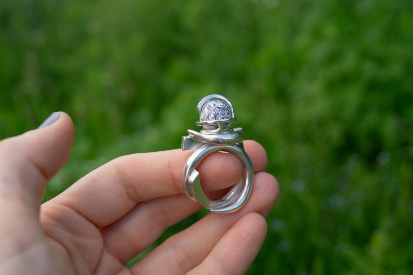 Photo of a person's hand holding a silver ring by Karoline Healy and H2ERG topped with a small orb above a grassy field