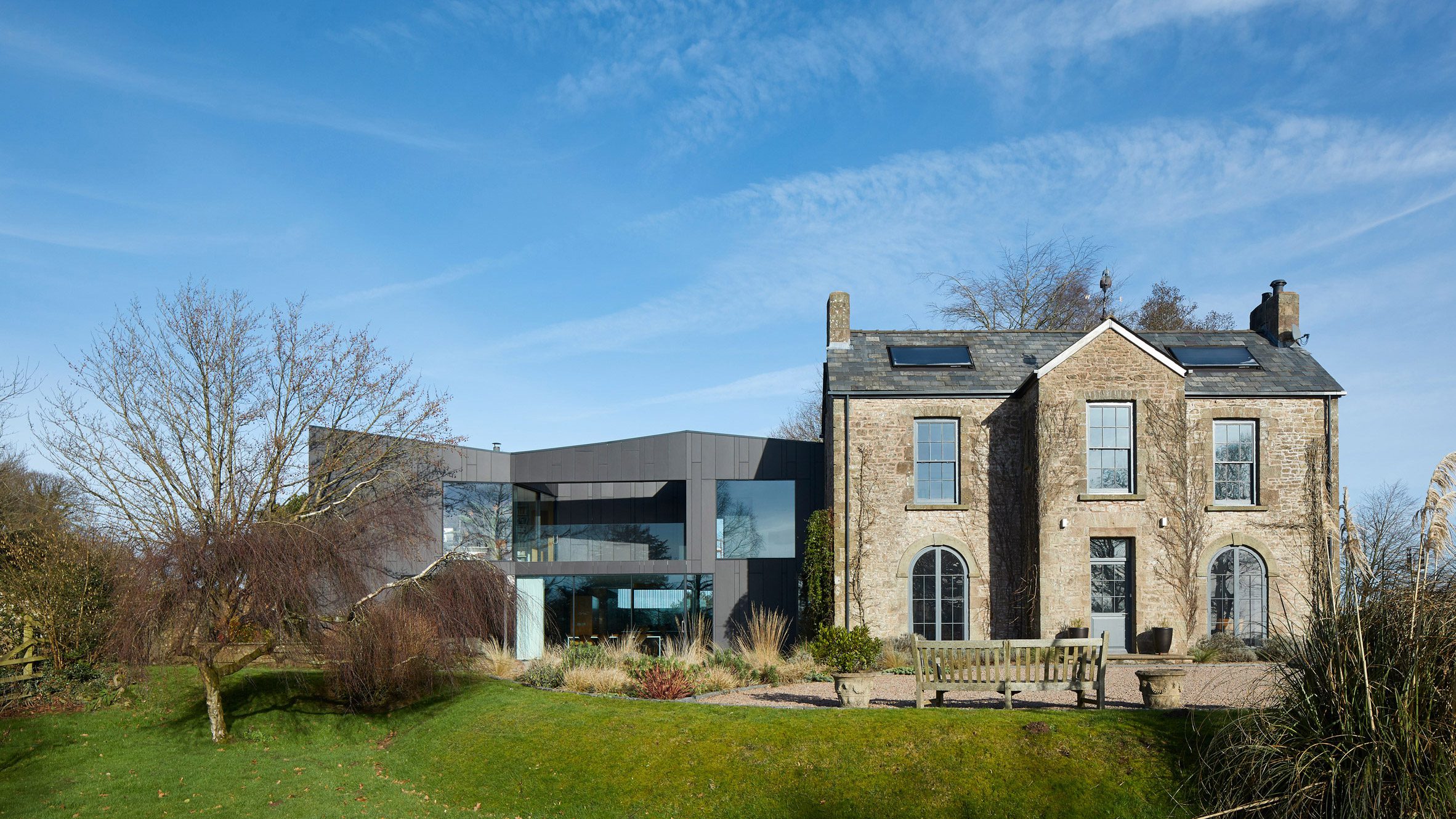 Photograph of a Georgian house in the countryside with a modern geometric extension. The extension is two-stories and has black cladding.