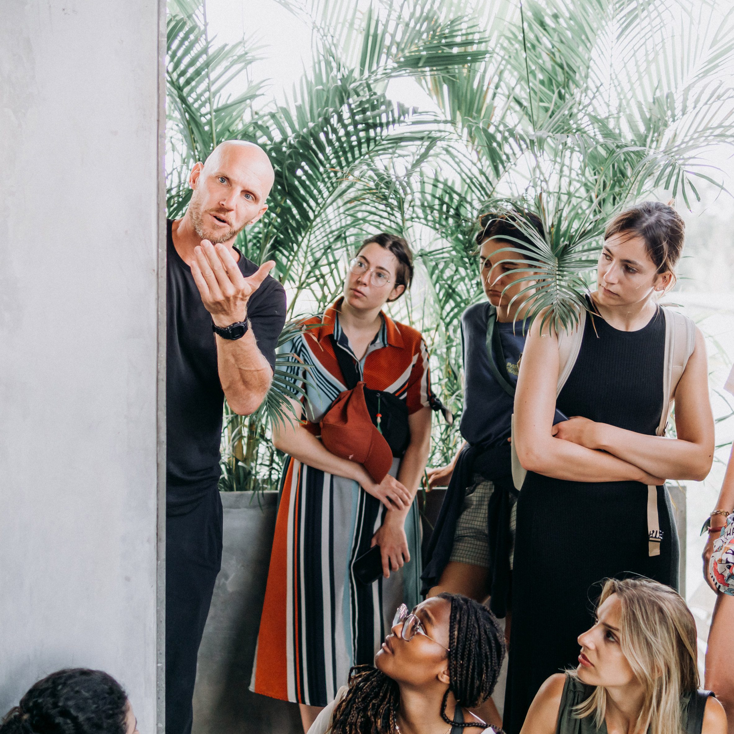 Photograph of a lecturer explaining work to a group of students in a classroom with a plant mural in the background.