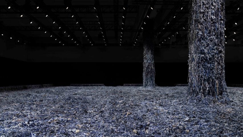 A photograph of a large floorspace covered in blue denim scraps, with two columns emerging from it, also covered. The columns lead to a black ceiling with multiple lights in it.