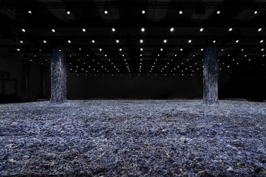 A photograph of a large floorspace covered in blue denim scraps, with two columns emerging from it, also covered. The columns lead to a black ceiling with multiple lights in it.