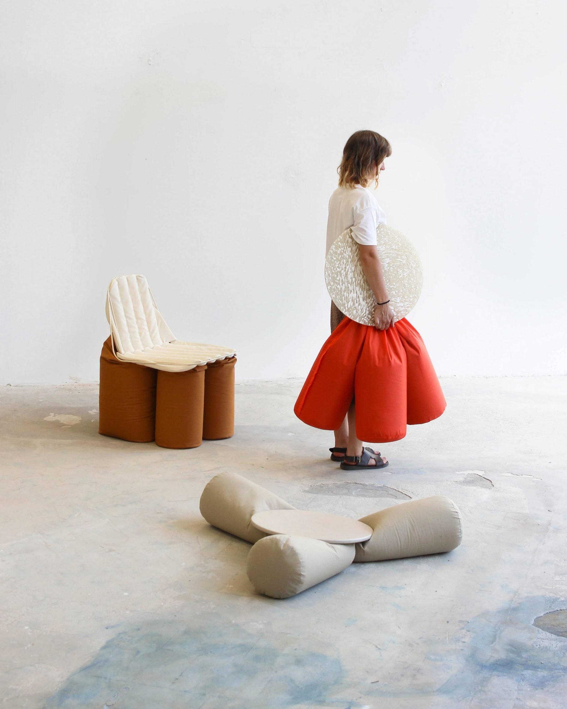 A photograph of a person, facing to the side, holding a seat featuring an orange cushion and a beige circular surface. There are two other seats surrounding them in tones of grey and brown.