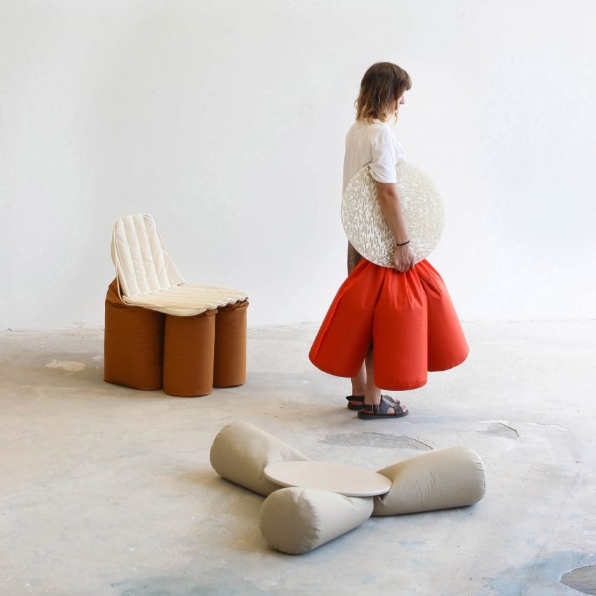 A photograph of a person, facing to the side, holding a seat featuring an orange cushion and a beige circular surface. There are two other seats surrounding them in tones of grey and brown.