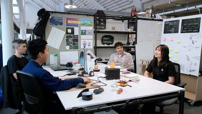 Photo of students Richard Alexandre, Karina Gunadi, Blake Goodwyn and Tanghao Yu sitting around a worktable surrounded by models and sketches for their Pyri device