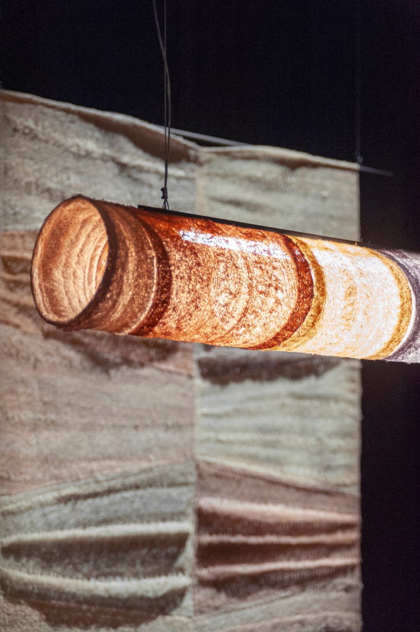 A photograph of a hanging illuminated pendant light in tones of orange and yellow against a backdrop of beige and black.