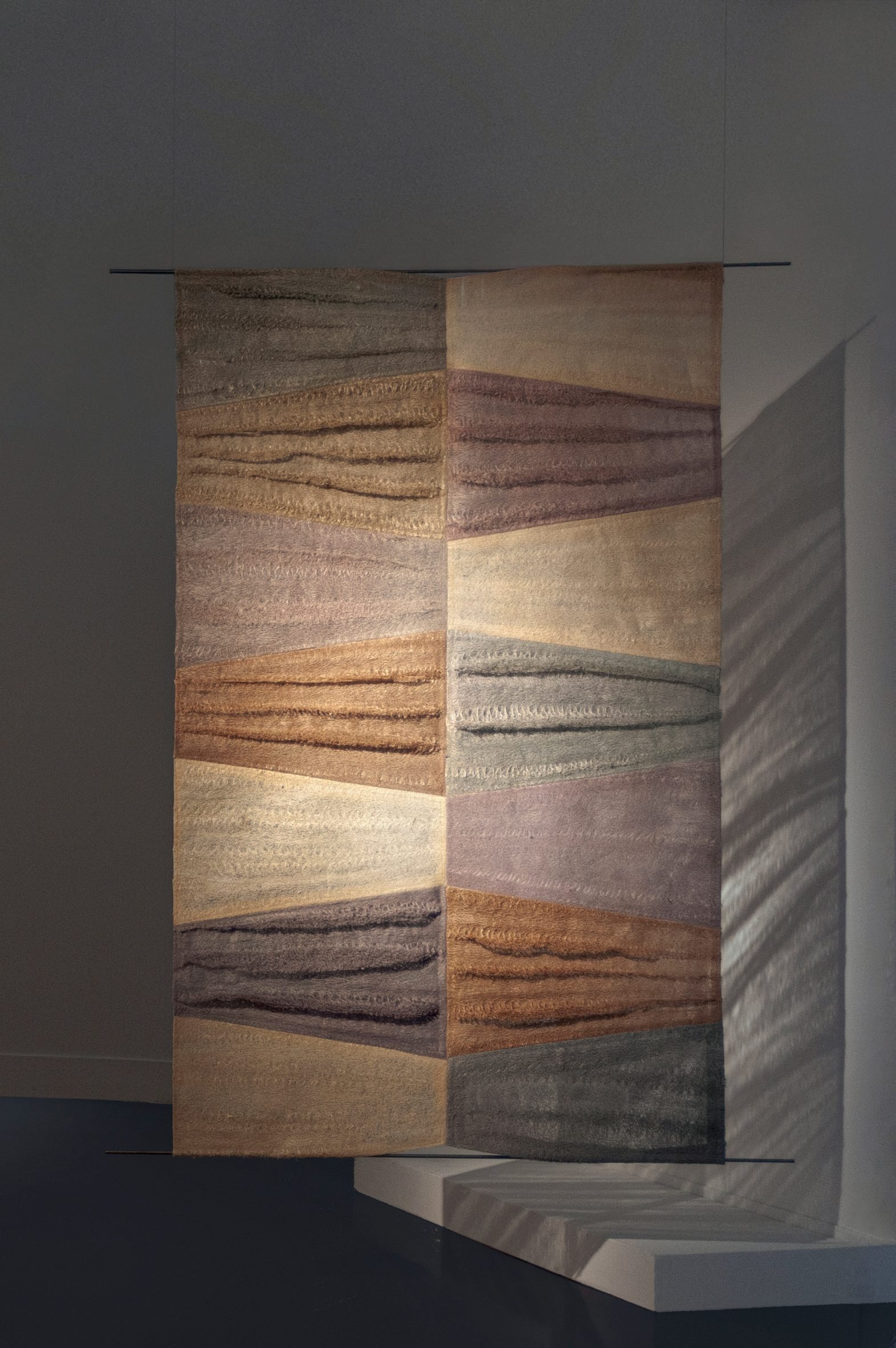 A photograph of a space divider with a geometric pattern on it in tones of beige, purple, blue and brown.