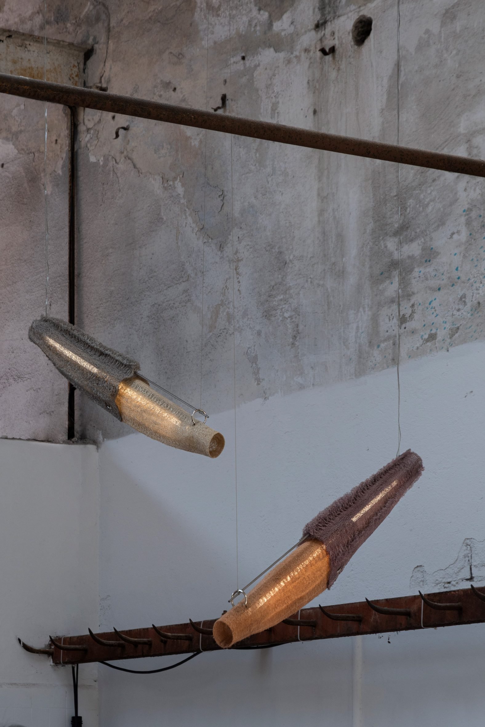 A photograph of two pendant lights, hanging diagonally, in tones of orange, beige and grey, against a white and grey concrete wall.