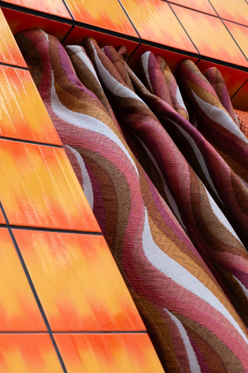 A photograph of a curtain in tones of pink, white, brown and orange, hanging from a structure consisting of orange tiles.