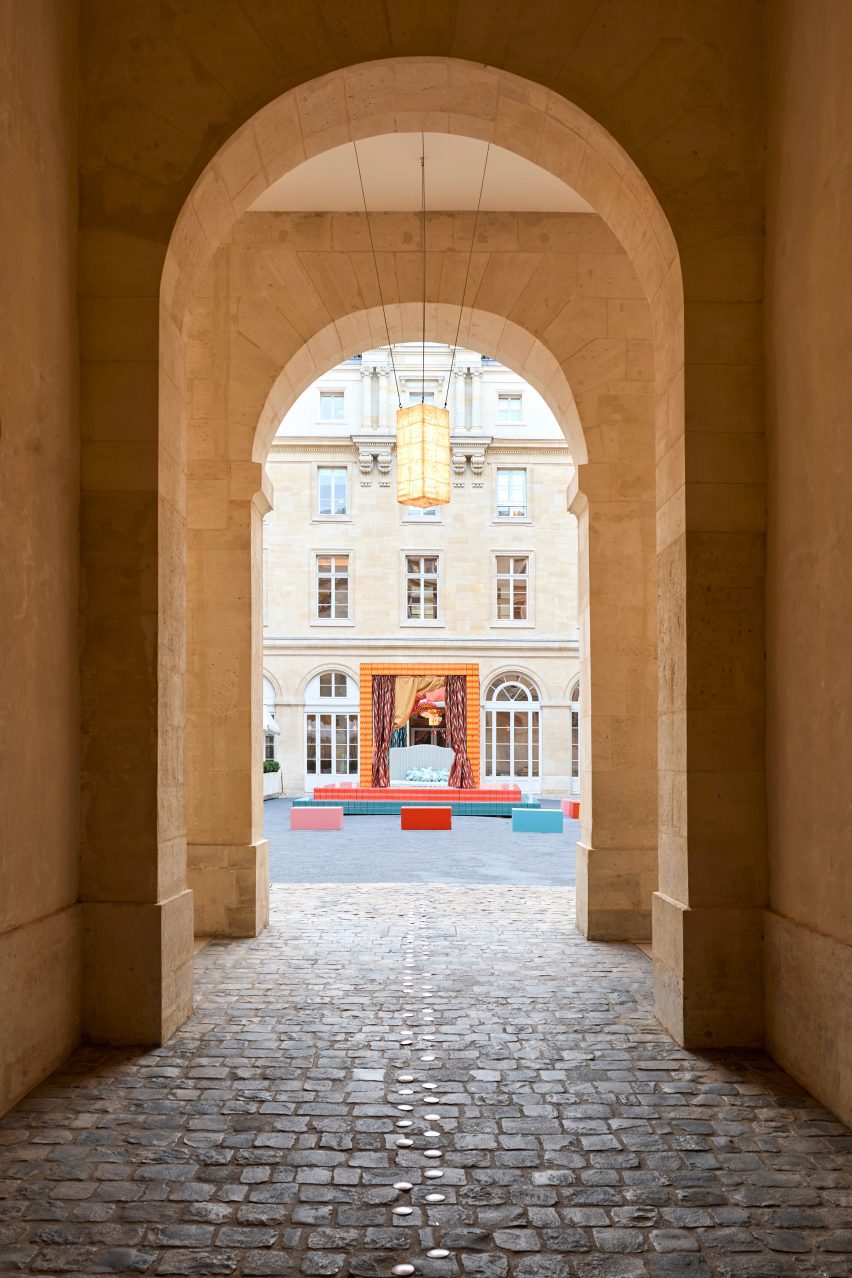 A photograph of an installation, captured through an archway, of a four poster bed in colours of blue, pink and orange, situated in a square of the Hôtel de la Marine.