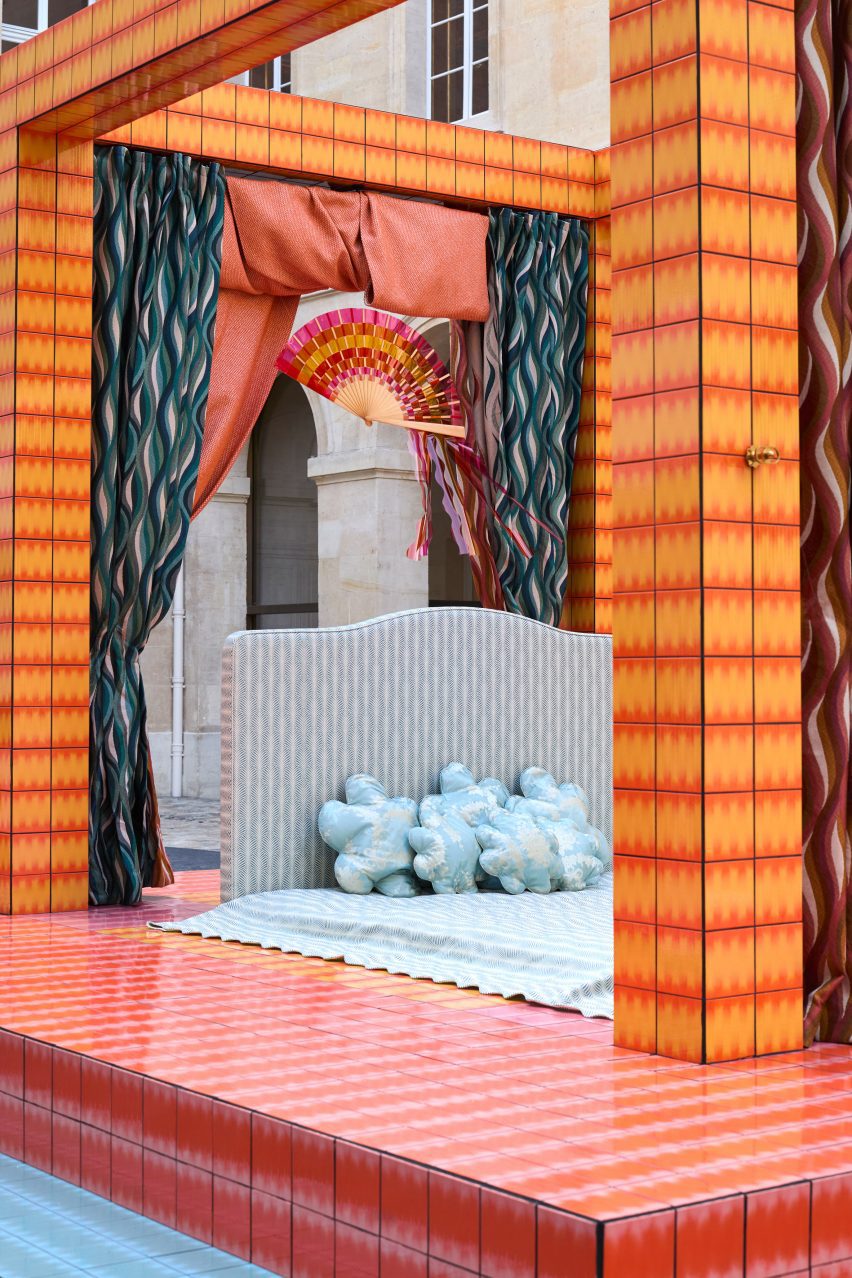 A cropped photograph of an installation depicting a four-poster bed in tones of orange, pink, red and blue.