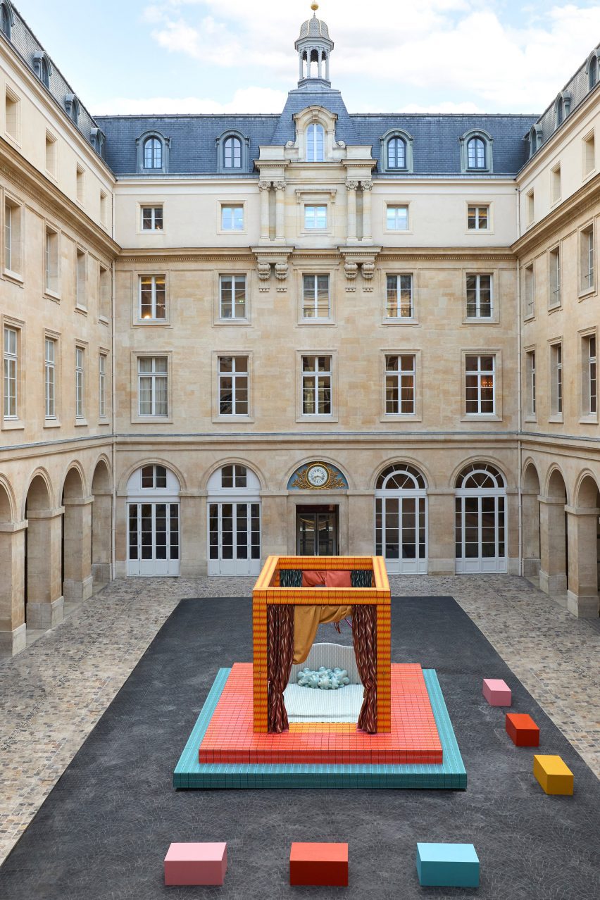 A photograph of an installation depicting a four-poster bed, in tones of orange, red, pink and blue, situated in a square in the Hôtel de la Marine.