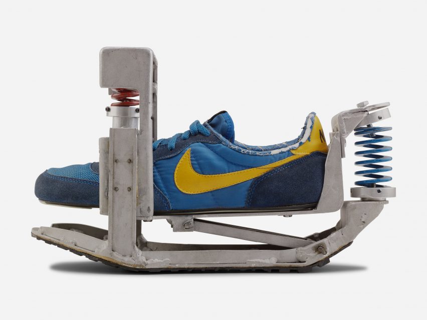 A photograph of a blue and yellow shoe prototype within a silver mechanical structure, against a white backdrop.