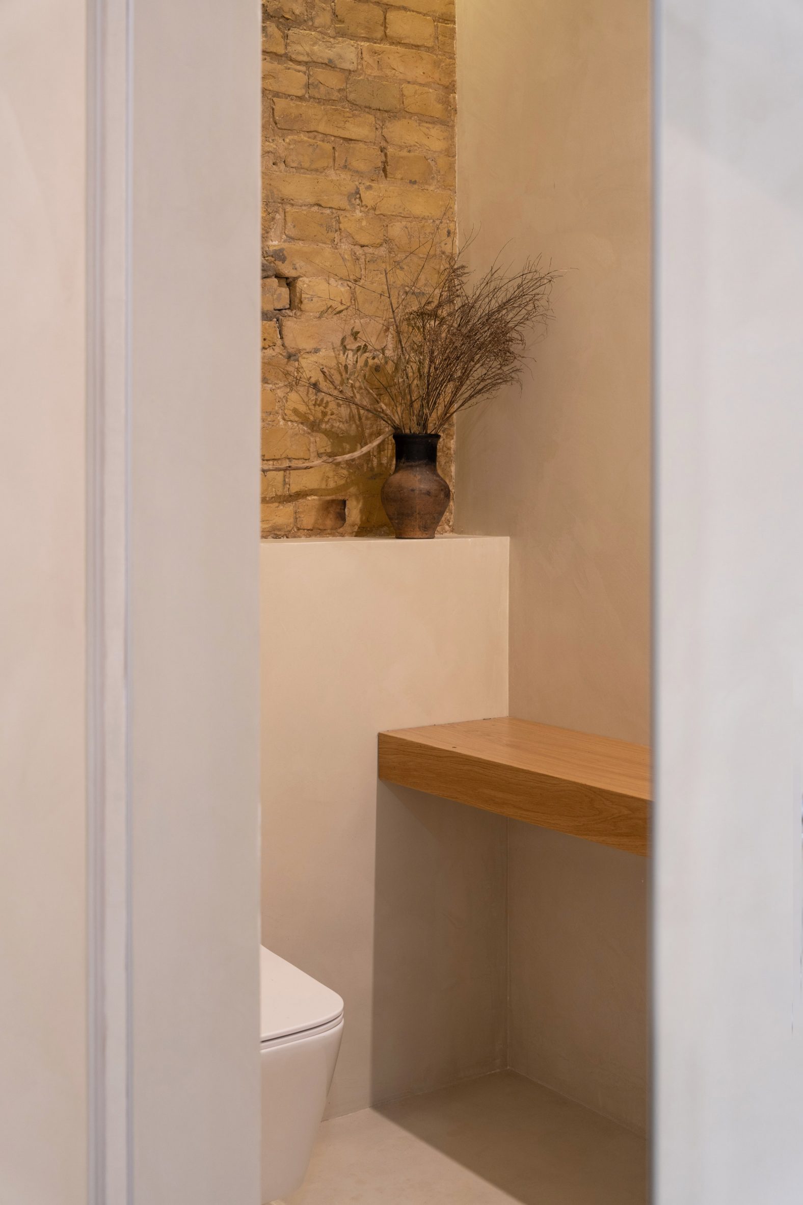 Photograph of a bathroom showing exposed yellow brick walls, a wooden shelf and a vase with flowers.