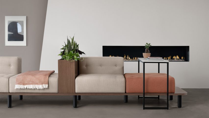 A photograph of a cushioned bench in tones of beige and orange with brown wooden features, positioned in front of a fireplace within a white wall. There is also a small table over the bench.