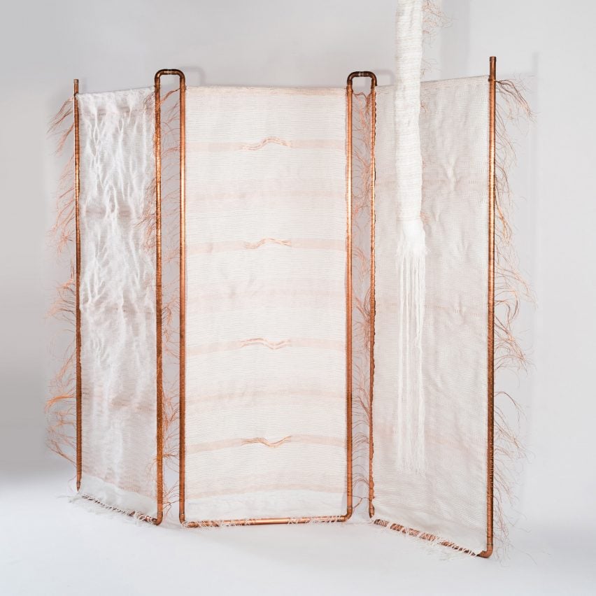 A photograph of a room divider, constructed of white fabric and a orange-toned copper frame. It is photographed in a white room.