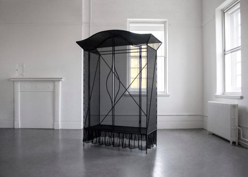 A photograph of a wardrobe, constructed with sheer black fabric, positioned in a white room with a grey floor. 