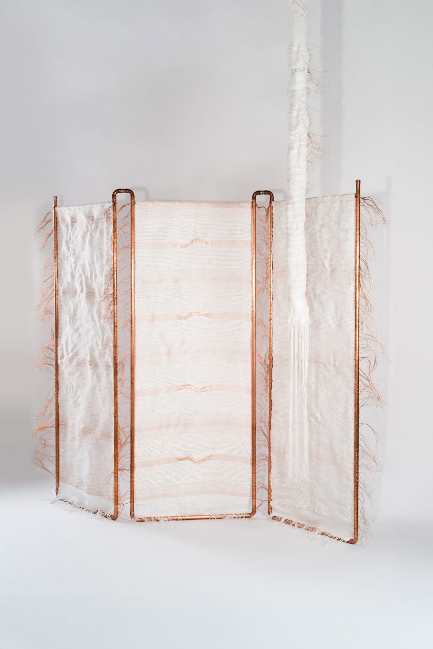 A photograph of a room divider, constructed of white fabric and a orange-toned copper frame. It is photographed in a white room.