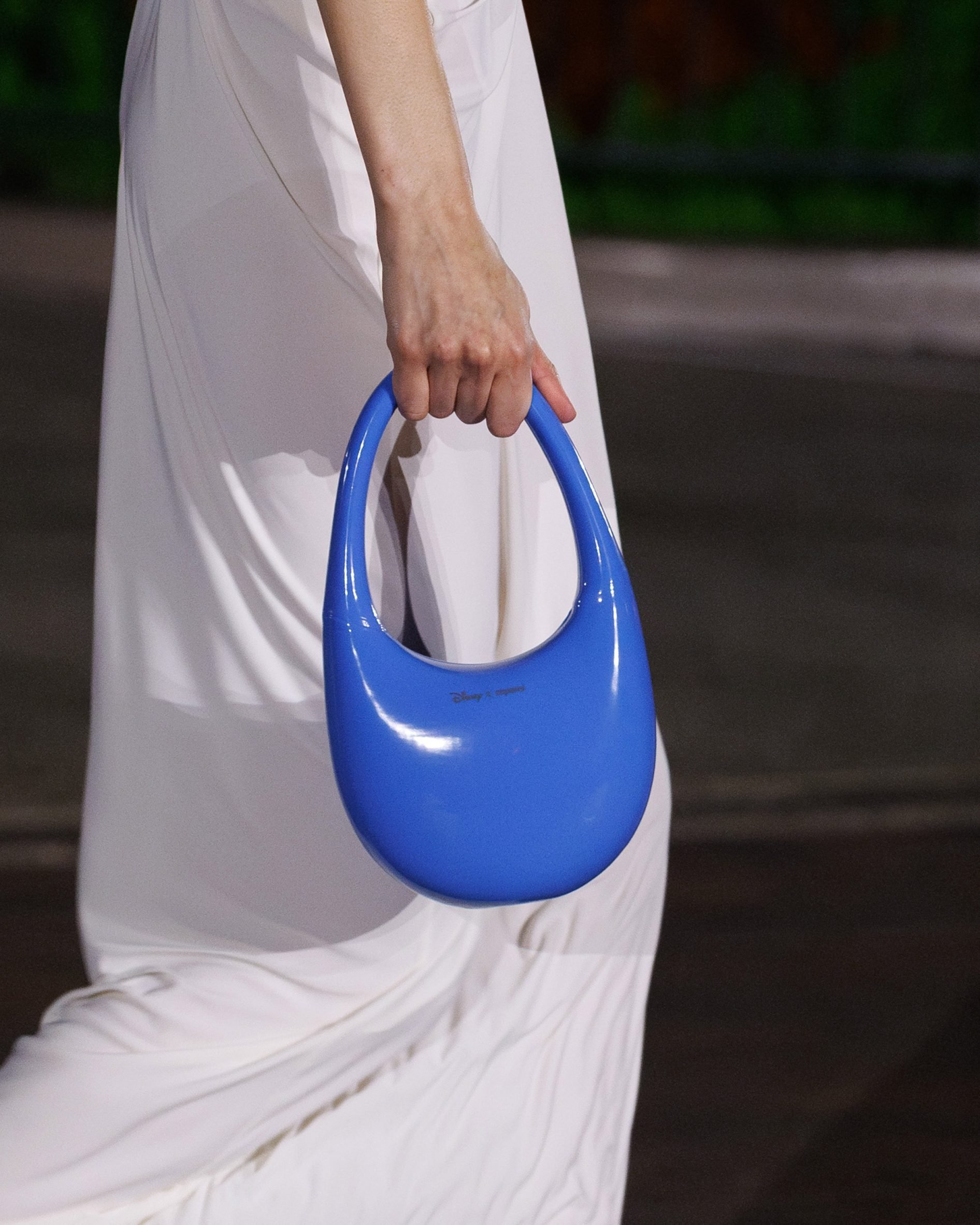 A close up photograph of a model's lower body on a runway wearing a white dress and shoes, carrying a blue handbag.