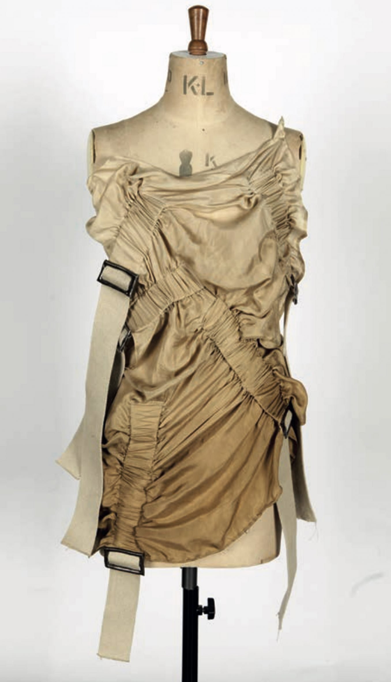 A photograph of a mannequin wearing a beige coloured garment with black buckles on it, against a white backdrop.