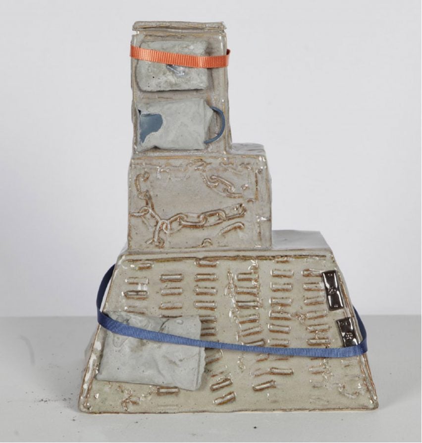 A photograph of a geometric ceramic sculpture in tones of grey, against a white backdrop. There are blue and orange strips of fabric wrapped around it.