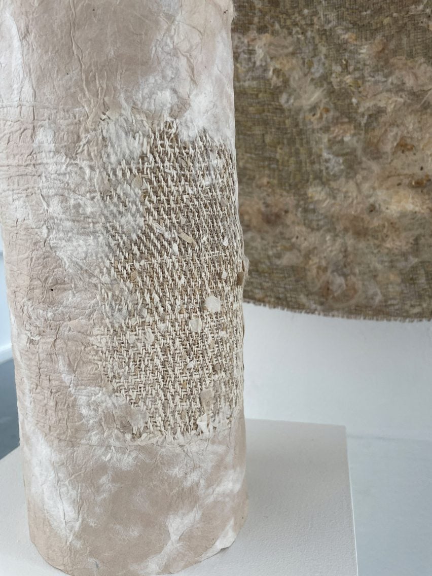 A photograph of a cylindrical object wrapped in a textile of beige tones. There is a brown textile sheet hanging behind it.