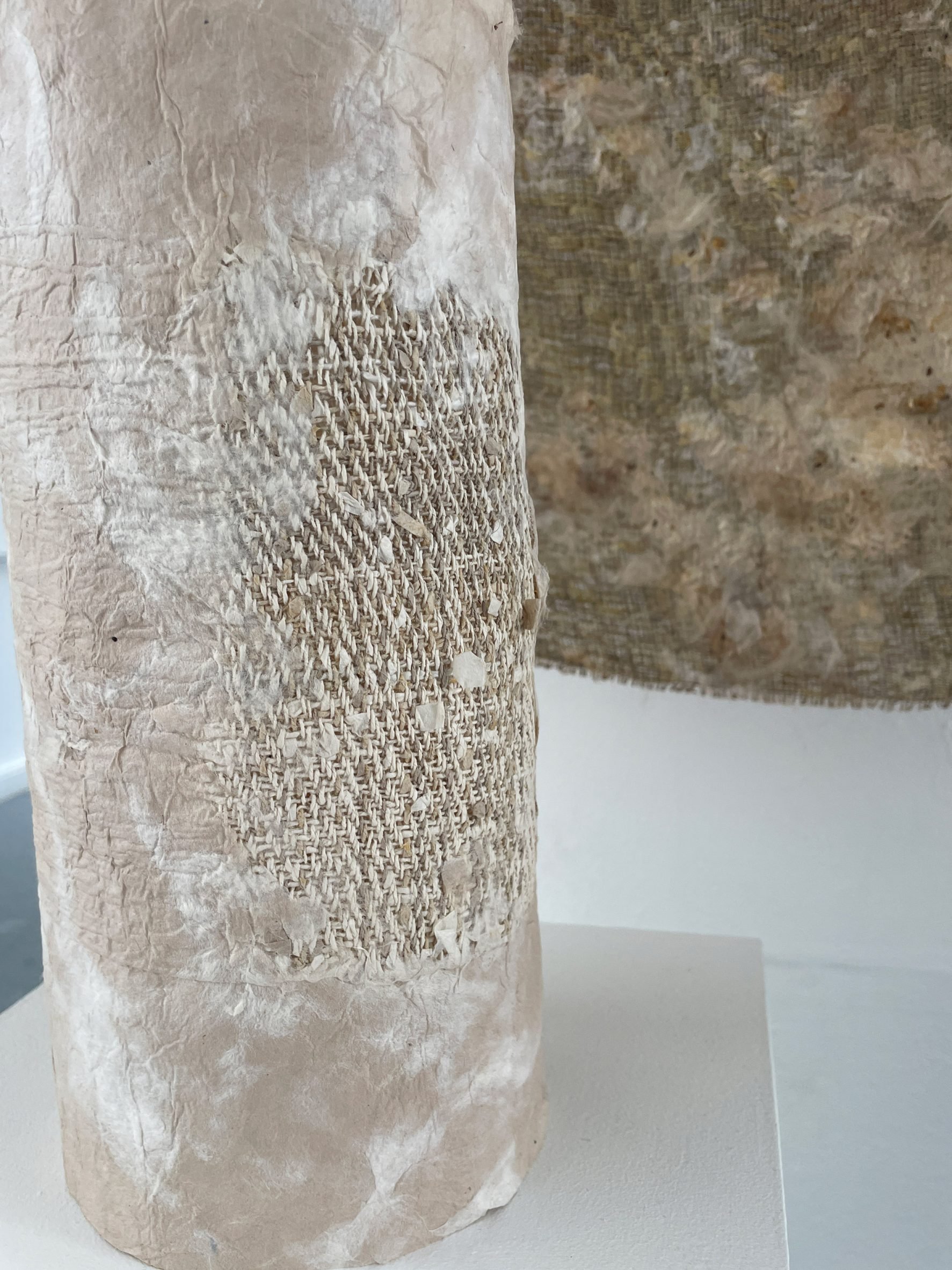 A photograph of a cylindrical object wrapped in a textile of beige tones. There is a brown textile sheet hanging behind it.