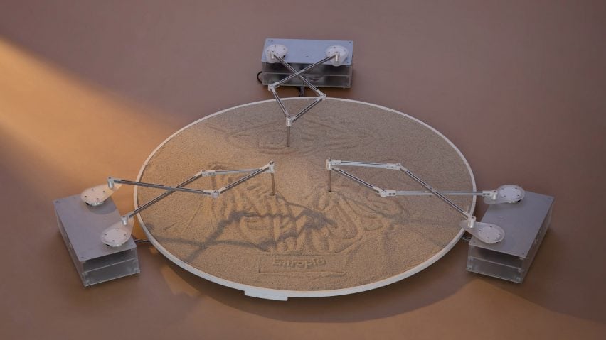 A photograph of an installation, with a circular vessel of sand and three drawing tools that are drawing patterns in the sand.