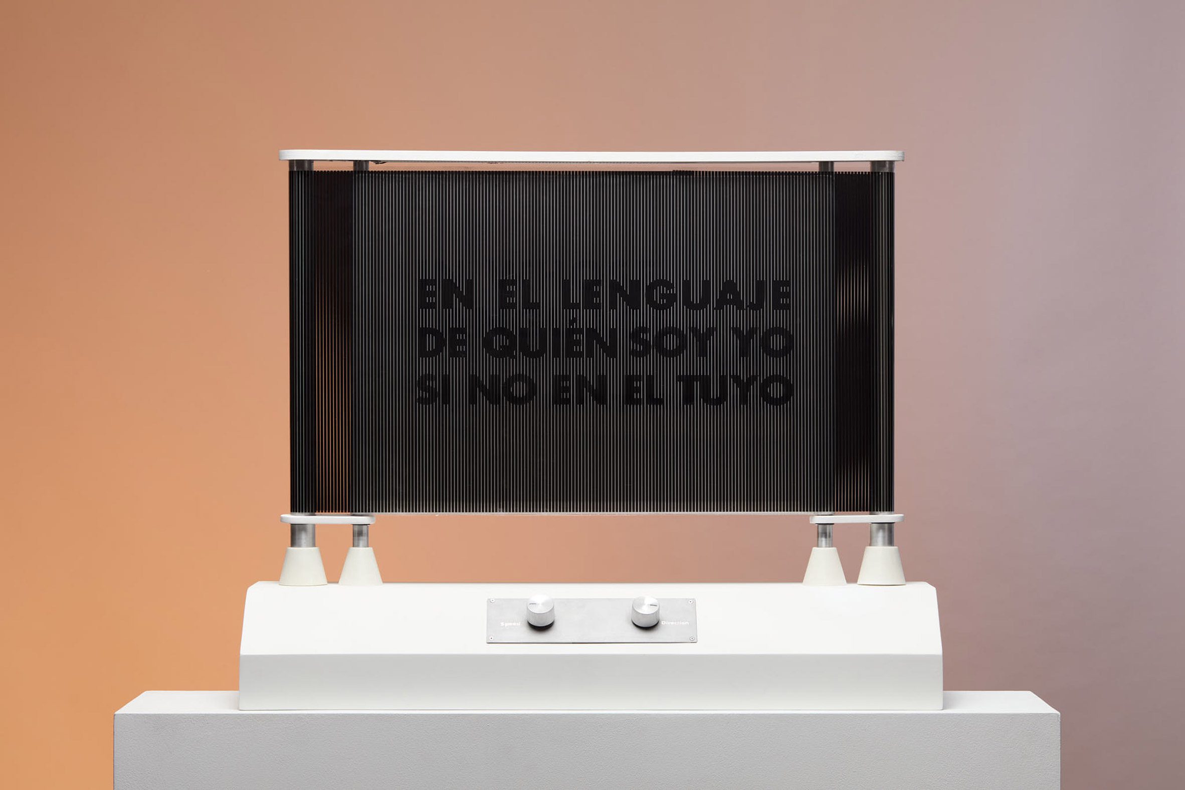 A photograph of an installation, with a white structure holding up a black screen, that says the words 'en el lenguaje de quién soy yo si no en el tuyo', in black text.