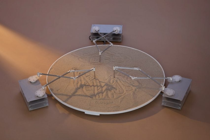 A photograph of an installation, with a circular vessel of sand and three drawing tools that are drawing patterns in the sand.