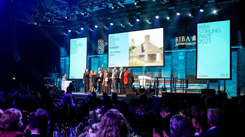 A photograph of an awards ceremony, showing people standing on a stage with screens behind them in tones of blue and white, and an audience of people sat in front of them.