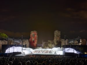 Wide photo of a bright white, angular open-air set full of performers under the night sky