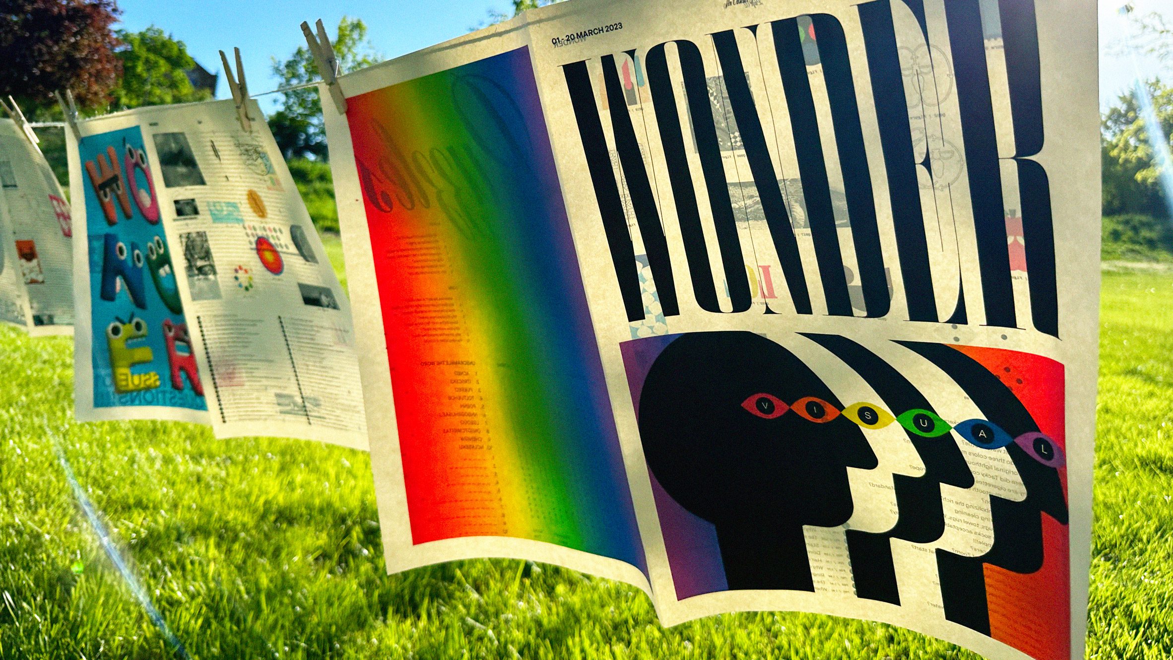 A photograph of sheets of newspaper hanging from pegs on a string line in a green space, with multicoloured details on them.