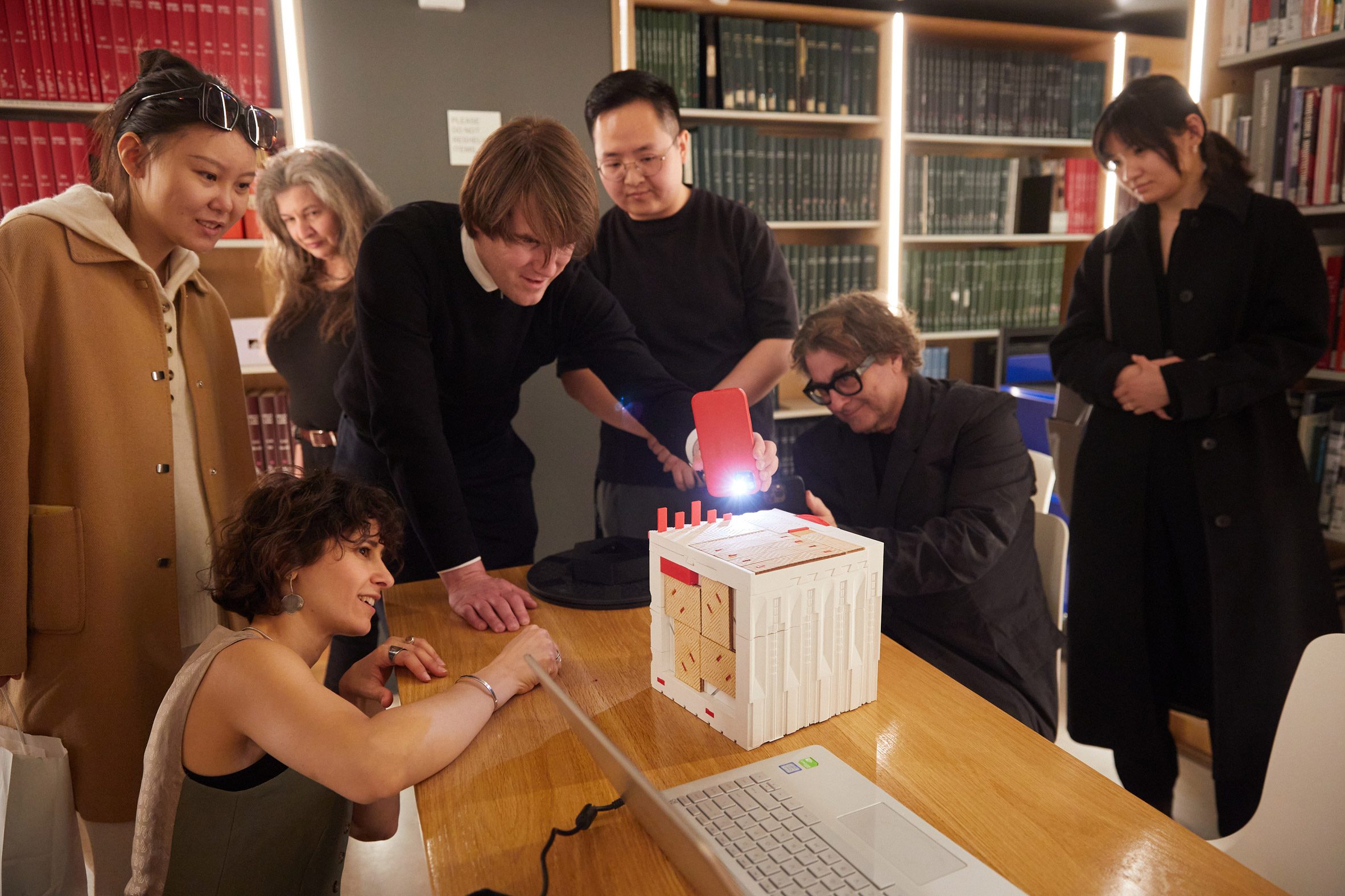 A photograph of a group of people around a table, interacting with a white cube.