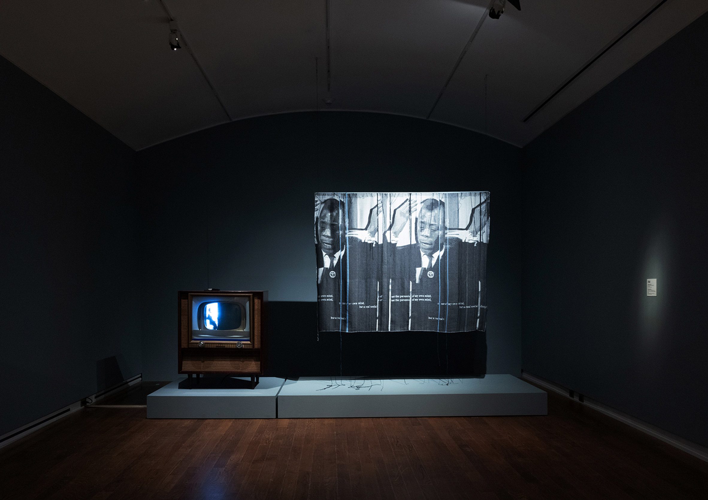 A photograph of an art installation, with a small television and curtain beside it, in tones of blue and and black. The installation is in a dark room with blue walls and a brown wooden floor.