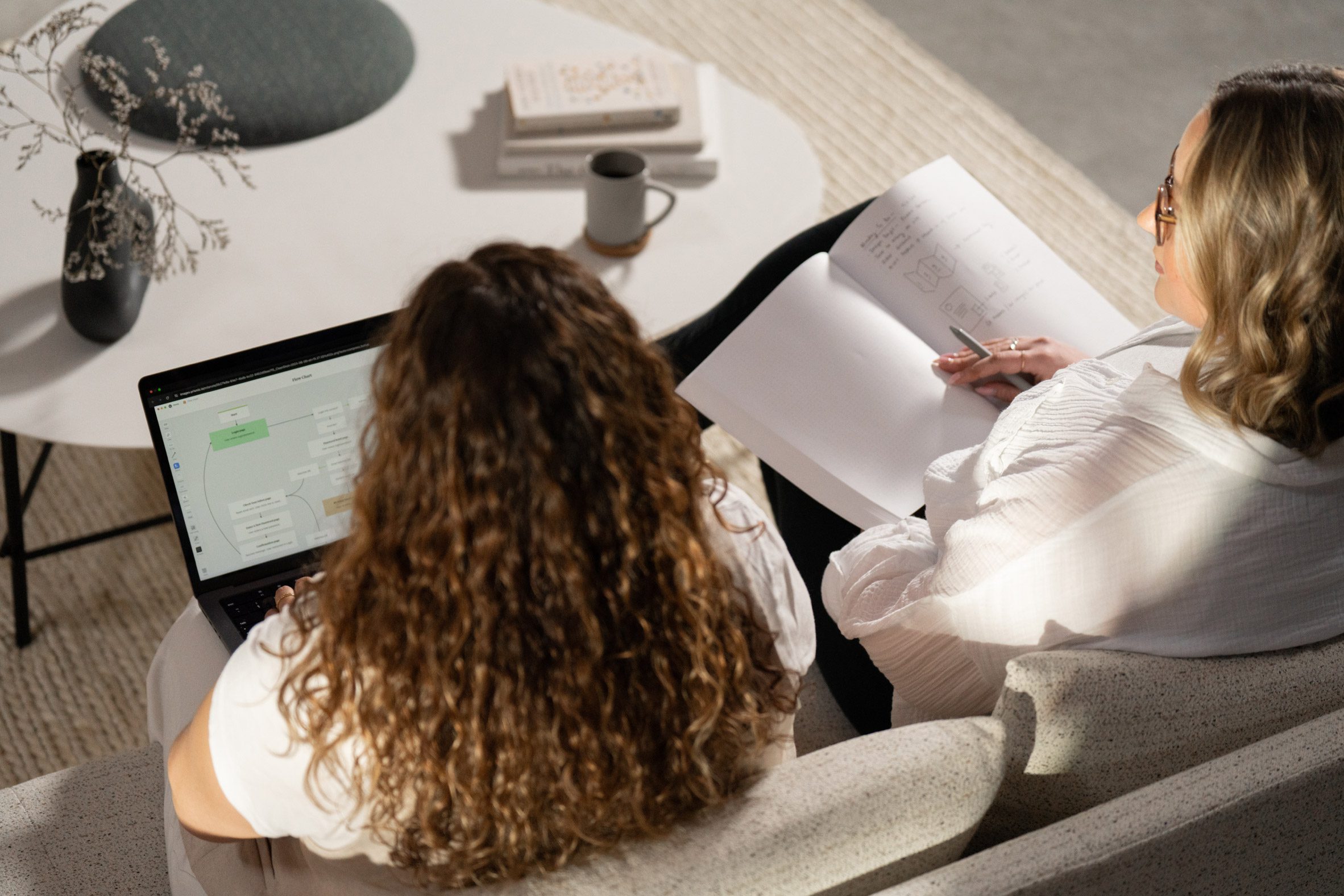 Photo of two women sitting on a sofa and collaborating on some work, seen from the back