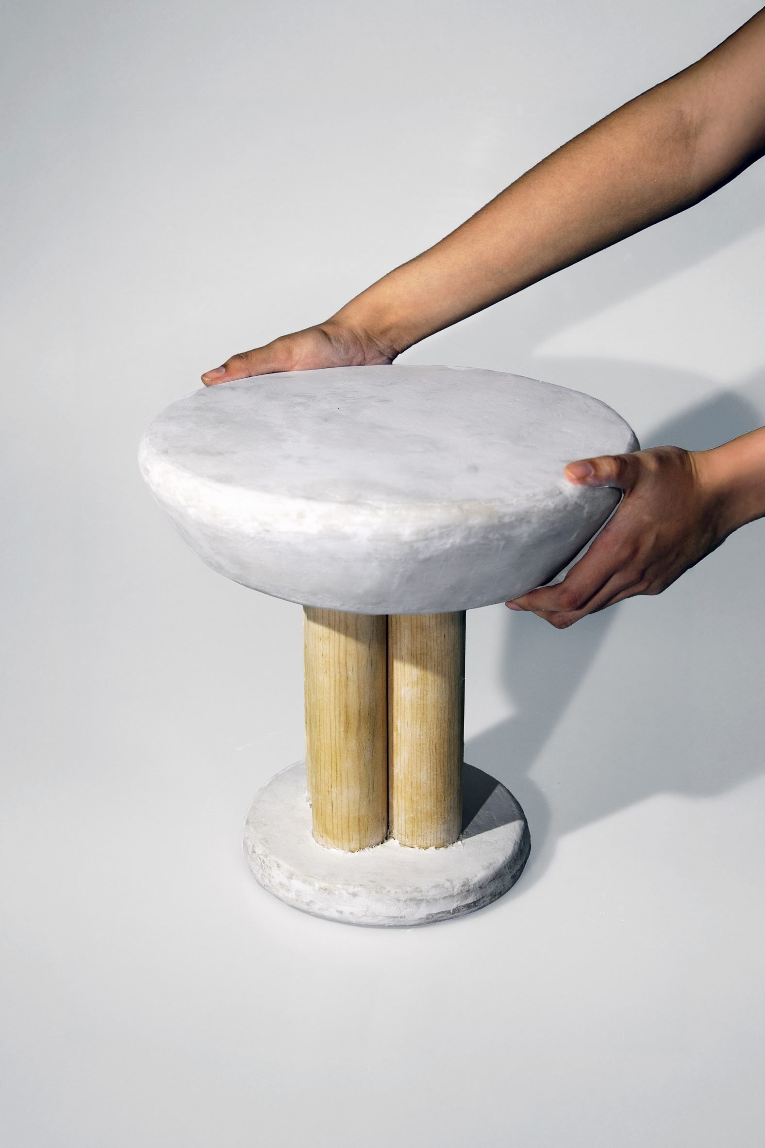 A photograph of a stool in tones of white, with a beige wooden leg. There is a person's hands interacting with it.