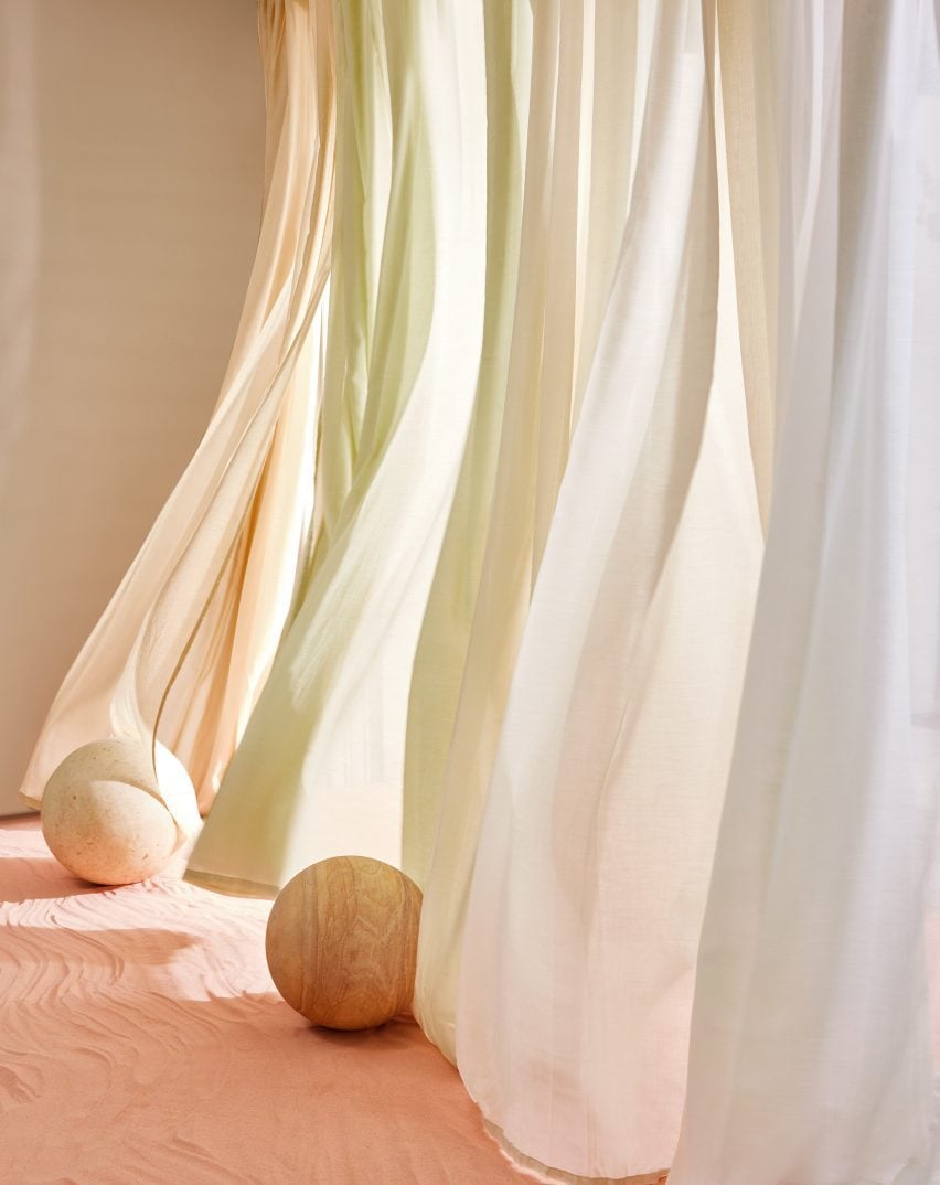 Photo of sheer curtains in pale colours hanging next to each other and blowing in the wind, along a minimally decorated corridor with a sand floor and two wooden spheres