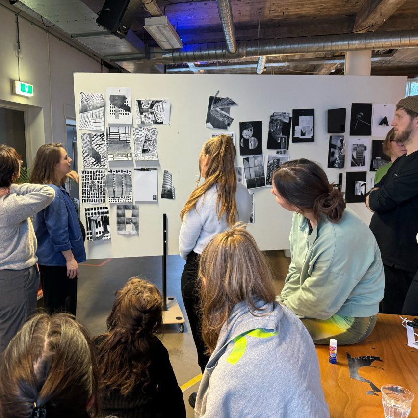 A photograph of students interacting, facing a board with various sheets of paper hung up on it.