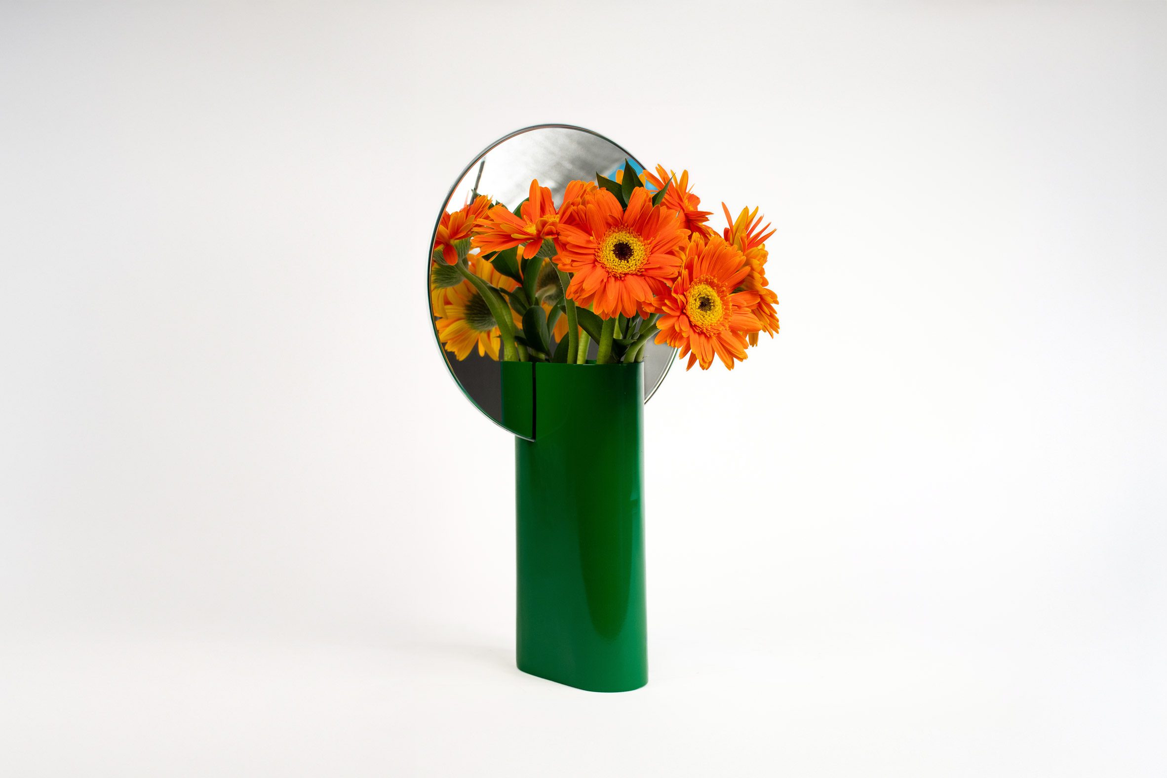 A photograph of a green vase with orange flowers inside, with a circular mirror integrated behind them. 