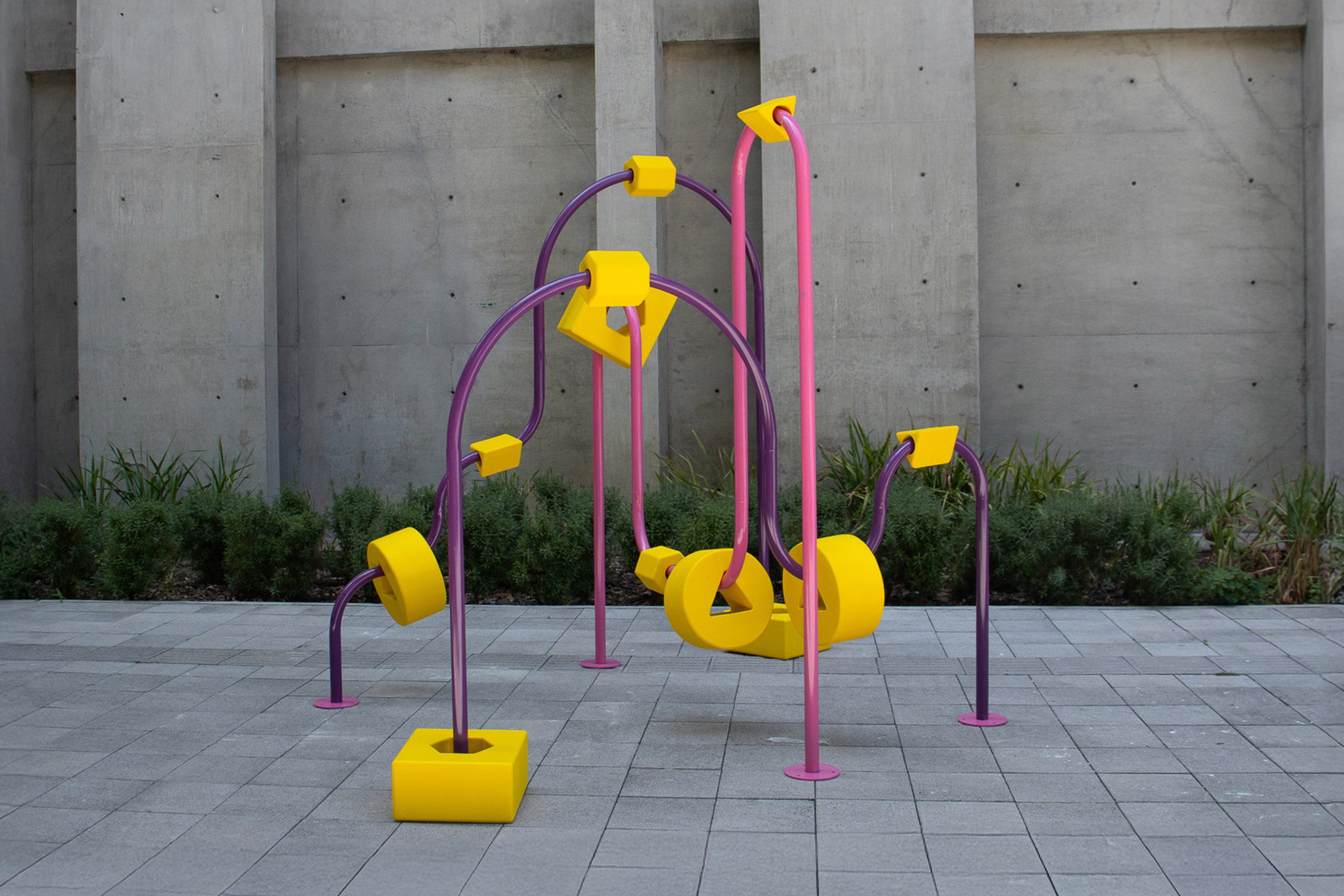 A photograph of an apparatus frame, in tones of pink and purple, within a grey concrete outdoor setting.