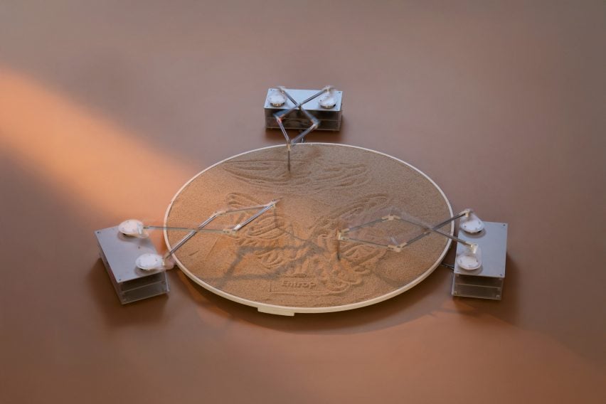 A photograph of an installation, in the form of a circular sand pit and three mechanic structures drawing forms into it.