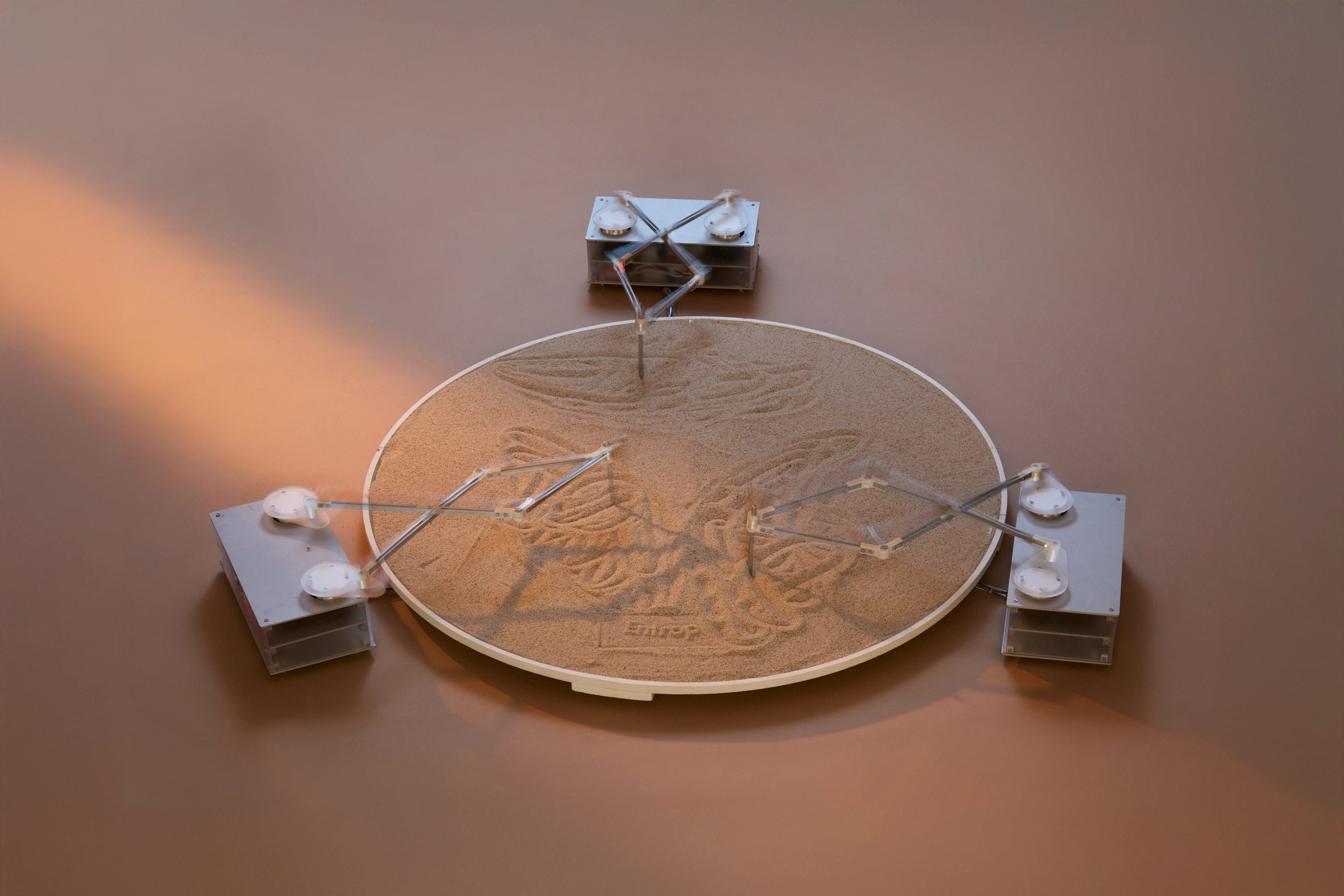 A photograph of an installation, in the form of a circular sand pit and three mechanic structures drawing forms into it.