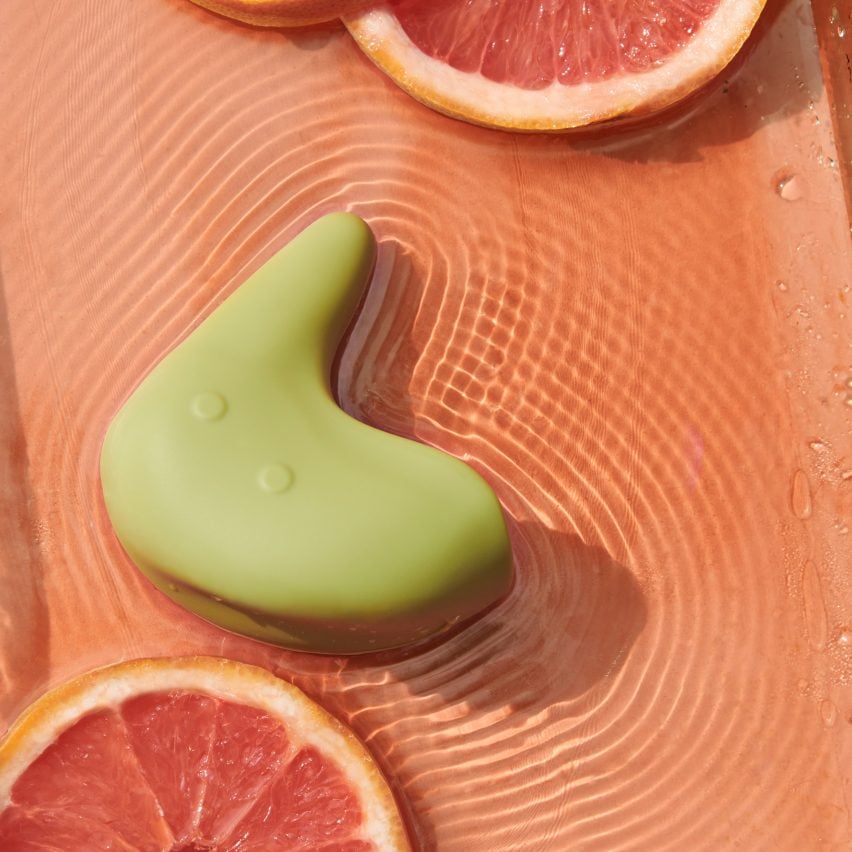 A photograph of a green vibrator which has an abstract, curved form, within a shallow body of pink water. There are slices of grapefruit in the water.