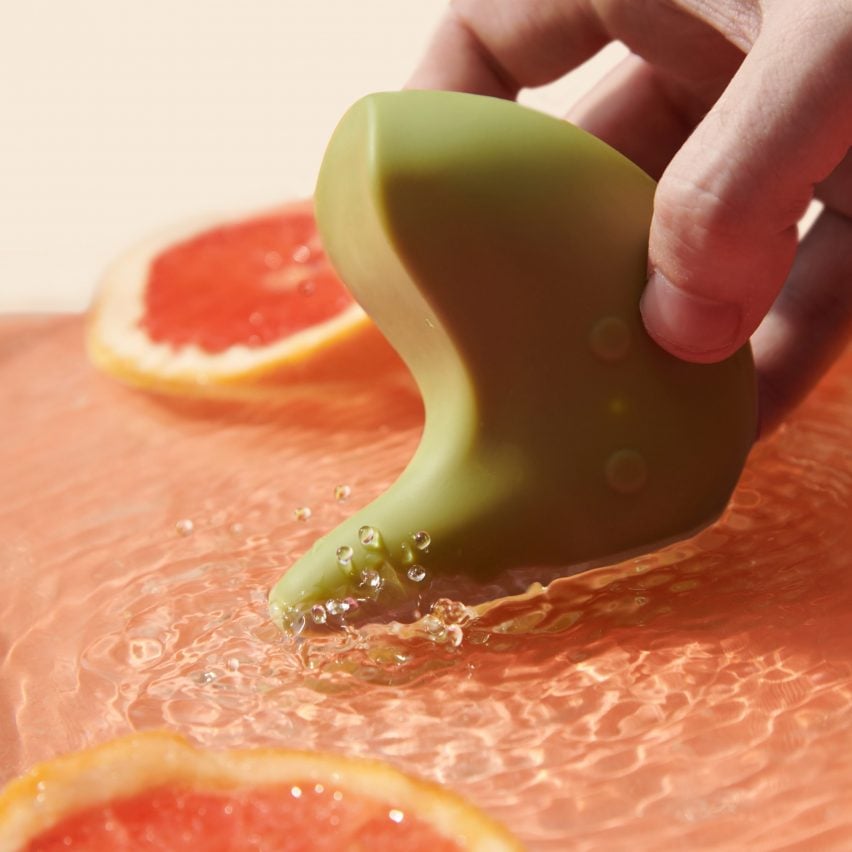 A photograph of a person's hand holding a green vibrator in an abstract, curved form, in shallow pink water. There are slices of grapefruit in the water.