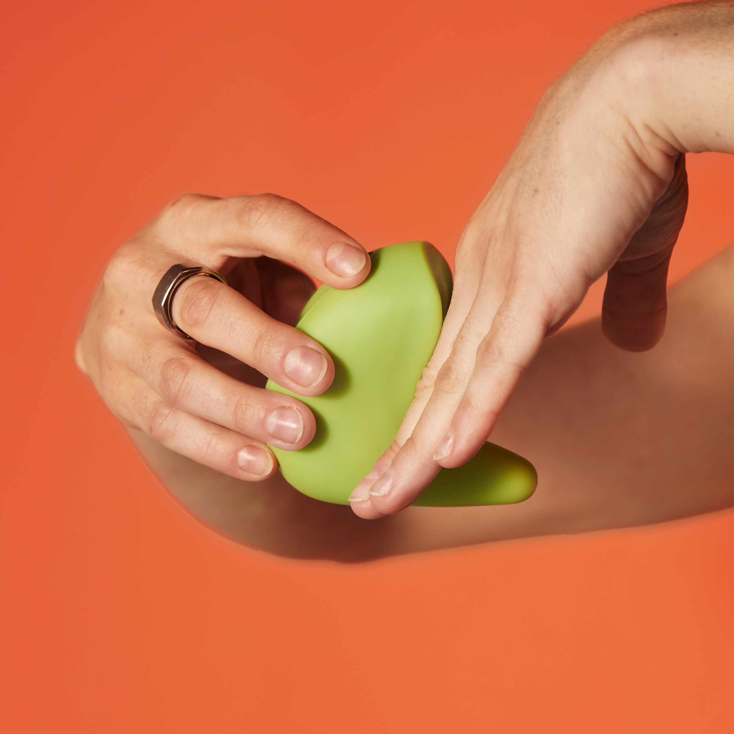 A photograph of a person's hands holding a green vibrator which has an abstract, curved form. There is an orange backdrop to the image.