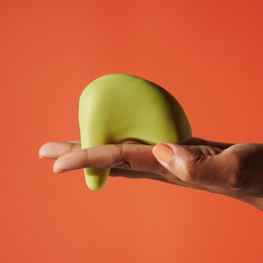 A photograph of a person's hand holding a green vibrator which has an abstract, curved form. The image has an orange backdrop.