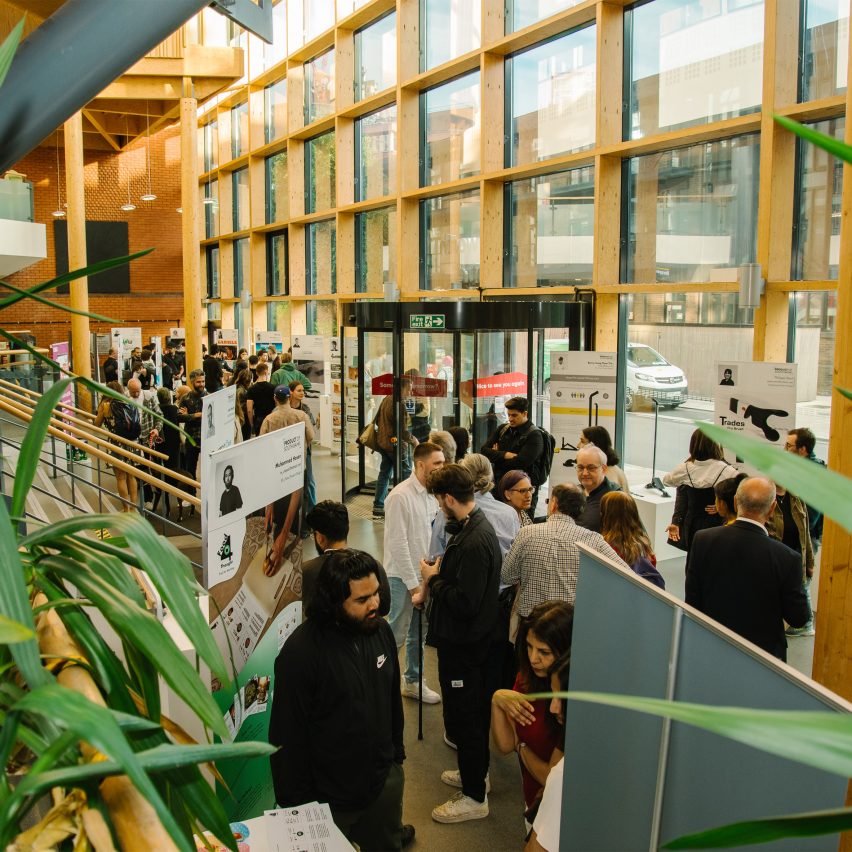 A photograph of a large room with a large group of people in it. There is a large glass wall on the right and there are green plants within the space.