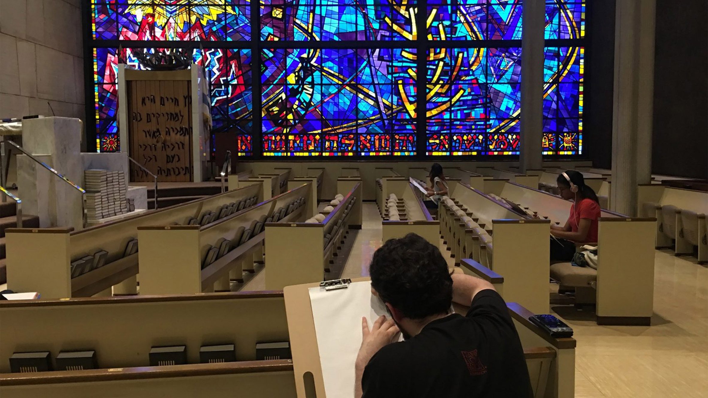 A photograph of people sat in a large room with multiple benches. There is a large, multi-coloured stained glass window in tones of blue, yellow, red and purple. One of the people is drawing the space.